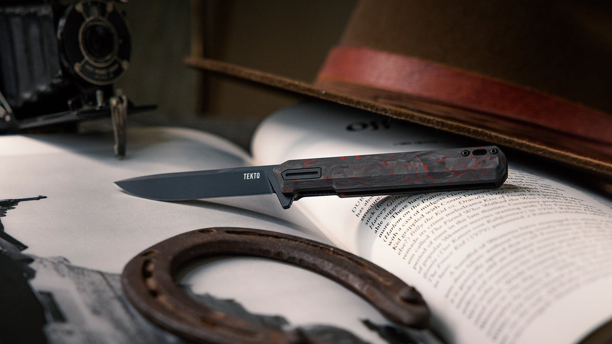 A TEKTO F2 Bravo folding knife resting on an open book beside a vintage camera and hat