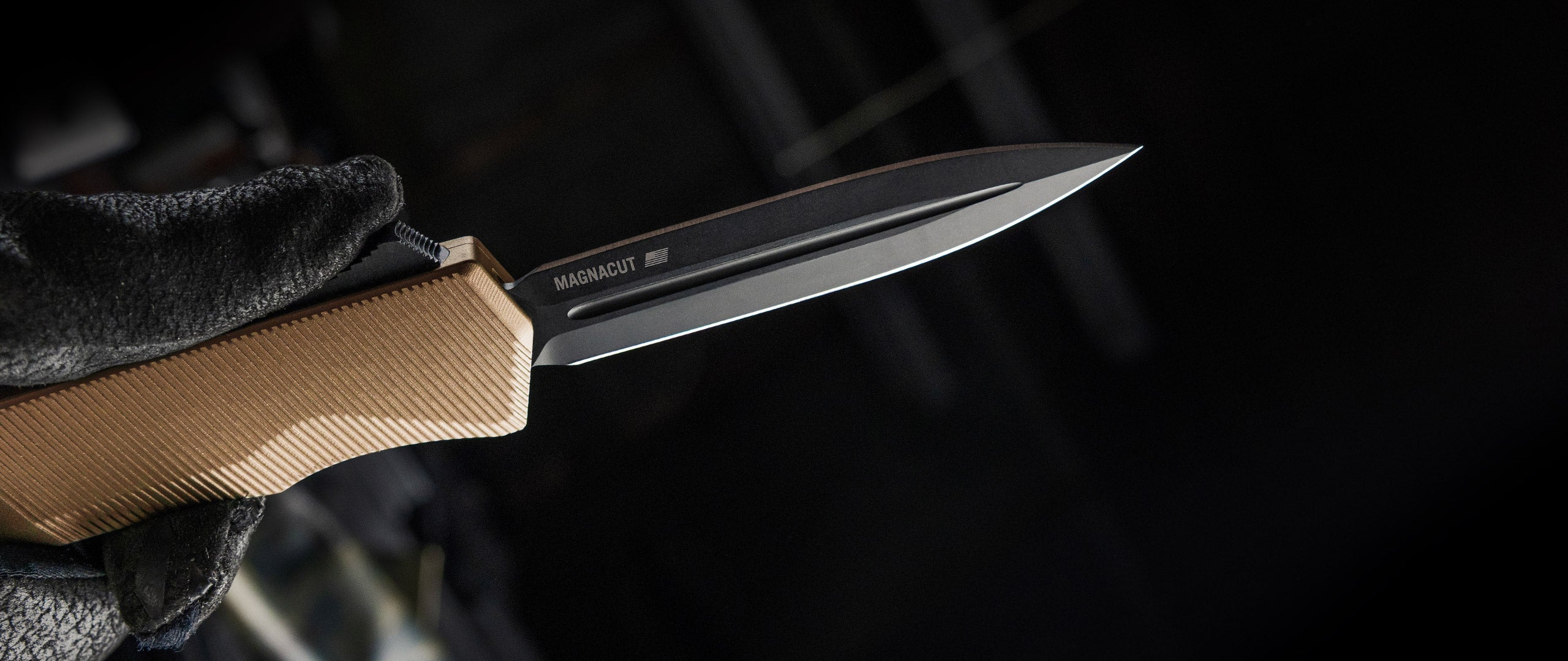 A close-up of a knife with a black blade and a tan textured handle, held by a black-gloved hand against a dark, blurred background. The blade is marked 'MAGNACUT'.