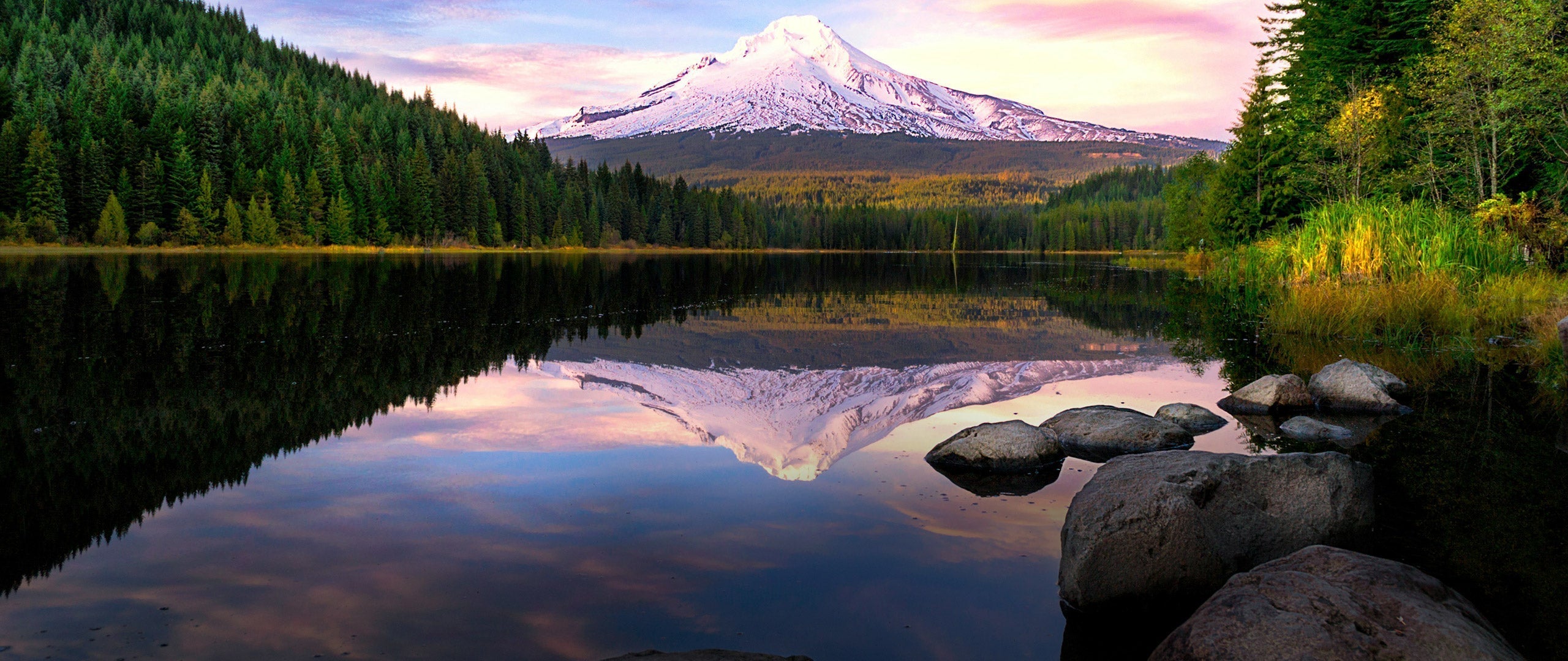 A scenic landscape photograph of a snow-capped mountain in Oregon reflected in a calm lake, surrounded by dense evergreen forests and large rocks in the foreground.