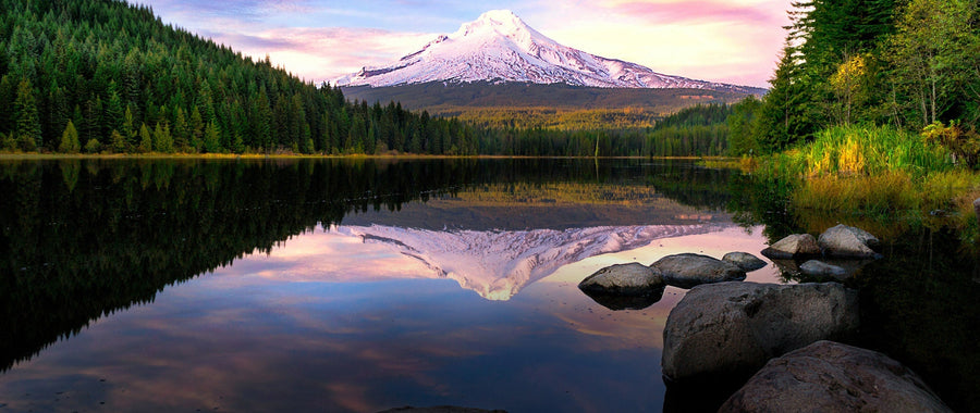 A scenic landscape photograph of a snow-capped mountain in Oregon reflected in a calm lake, surrounded by dense evergreen forests and large rocks in the foreground.