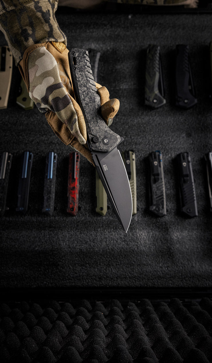 Hand holding a folding knife F3 Charlie from Tekto Knives above a collection of knives on a foam surface