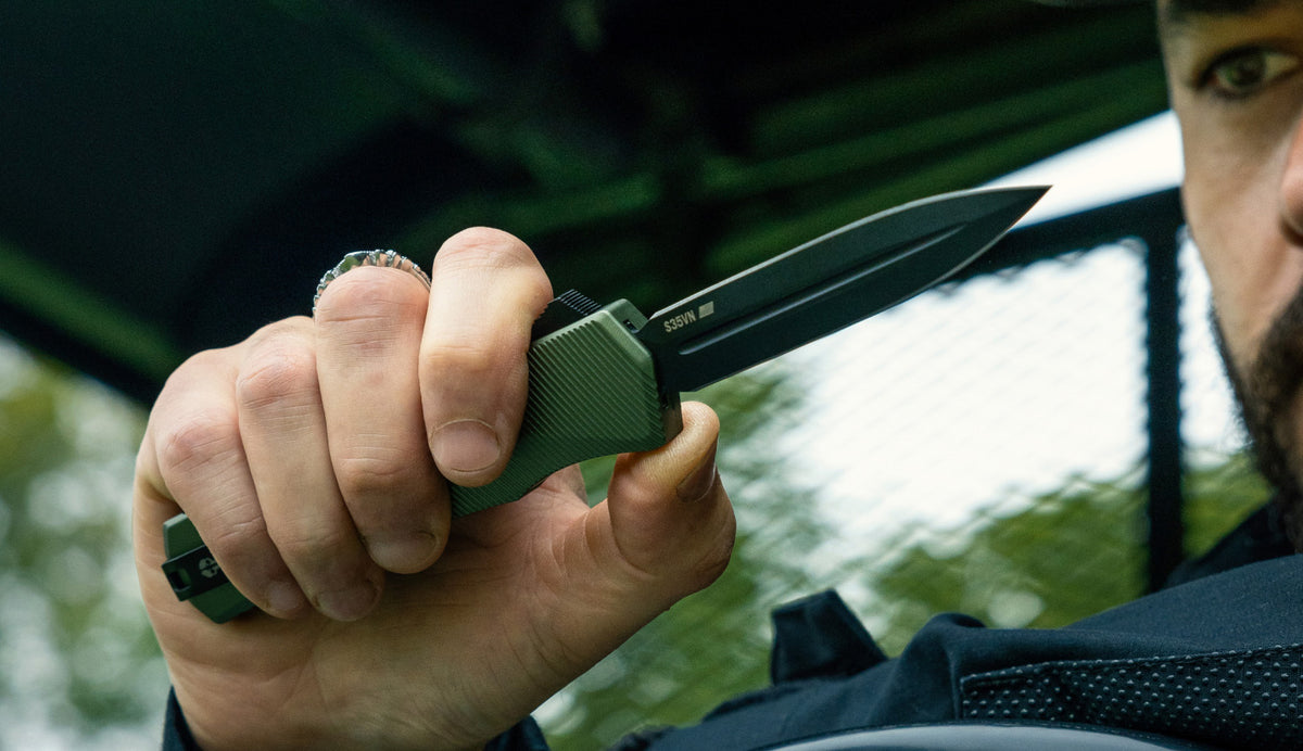 Hand holding a TEKTO OTF knife with an olive green handle and a double edge blade
